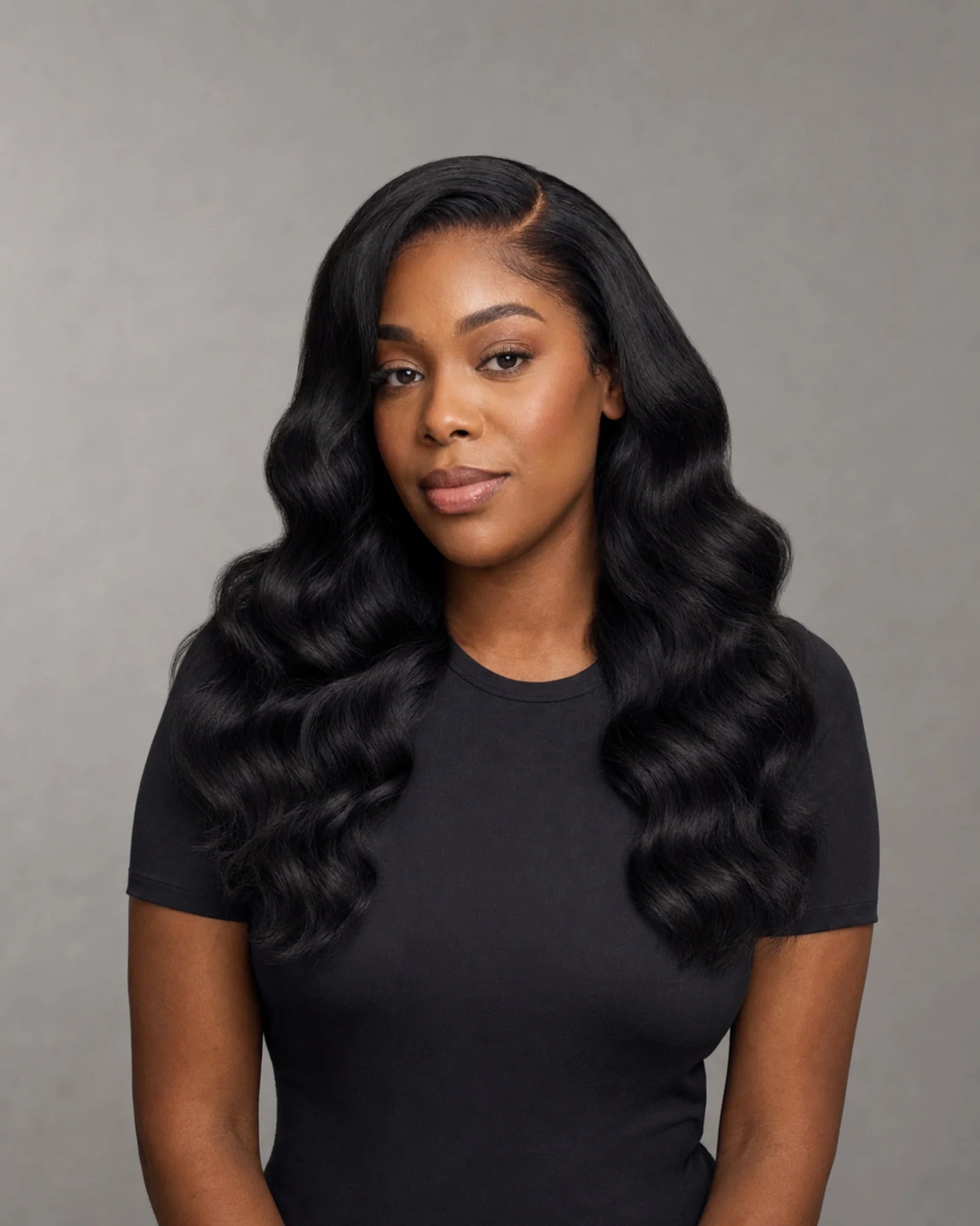Front view of a Black woman wearing a full natural-black loose-wave wig with a defined side part.