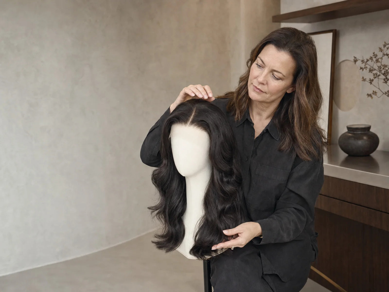 Woman performs a final visual check on a finished long wavy wig displayed on a mannequin head.