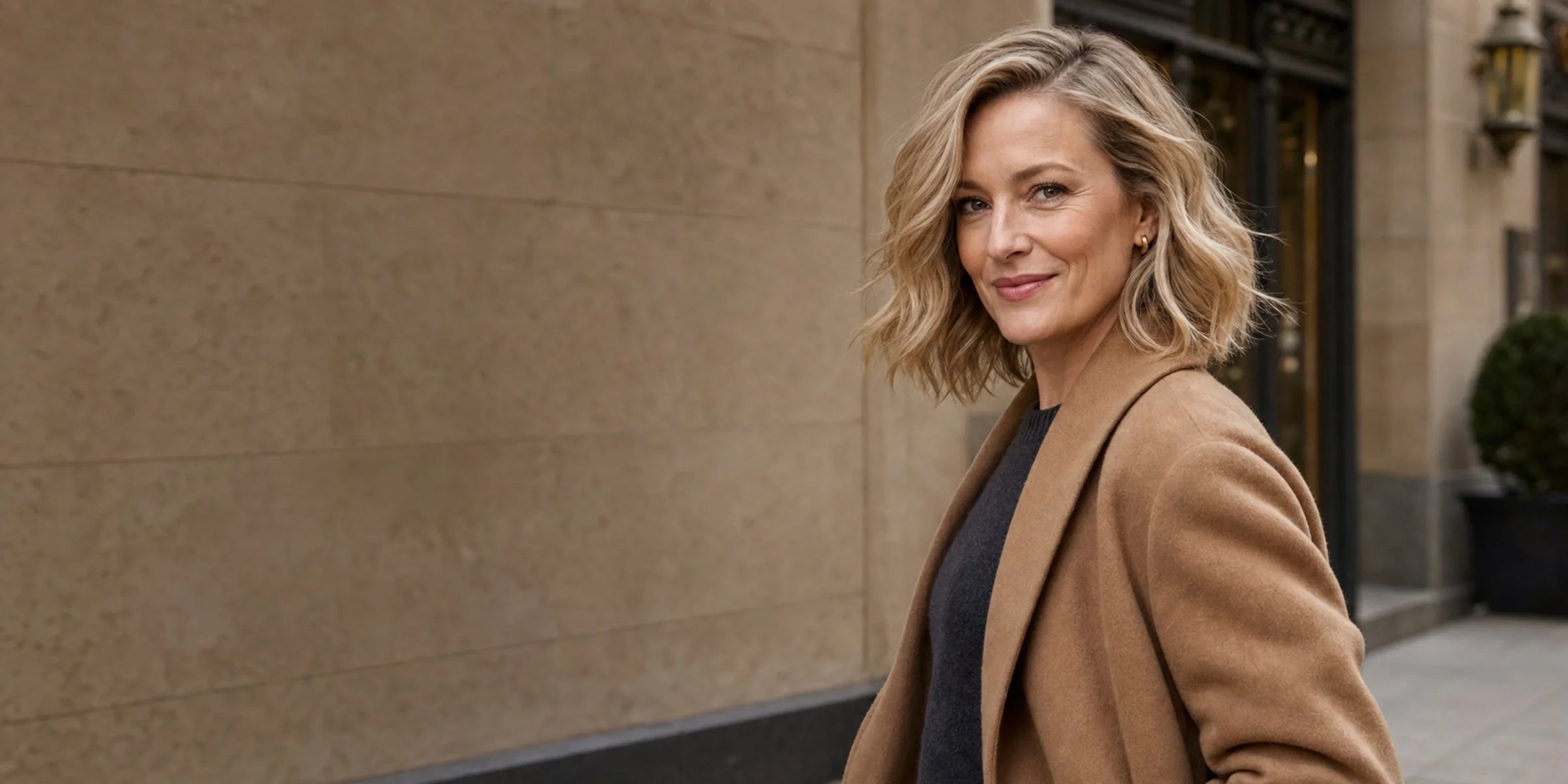 White woman in a camel coat wears a short rooted beige-blonde textured bob in a city setting.