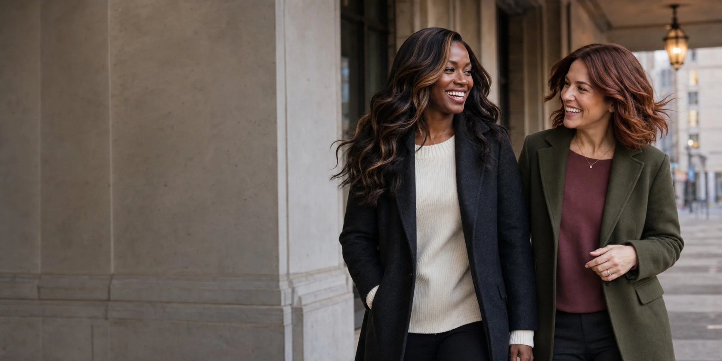 A Black woman in long highlighted waves walks beside a White woman with an auburn bob through a refined city setting.